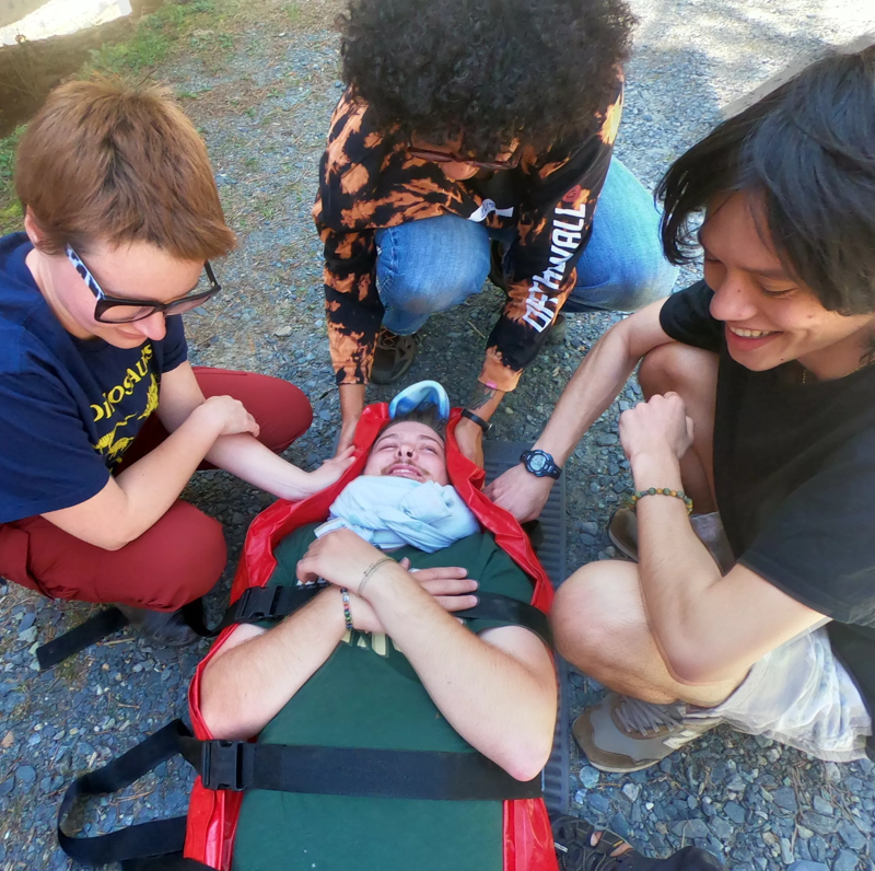 In the image, a person is lying on a red and black stretcher, secured with straps. Three individuals are attending to them. One person on the left is holding the person's arm, while another behind the stretcher is stabilizing their head. A third person on the right is smiling. The setting appears to be outdoors, with a gravel or paved surface visible beneath the stretcher.